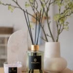 amber-and-sauge-200ml-fragranced-diffuser-alchemy-with-golden-collar-black-reeds-black-label-with-golden-foiled-branding-on-a-table-with-a-vase-in-the-background